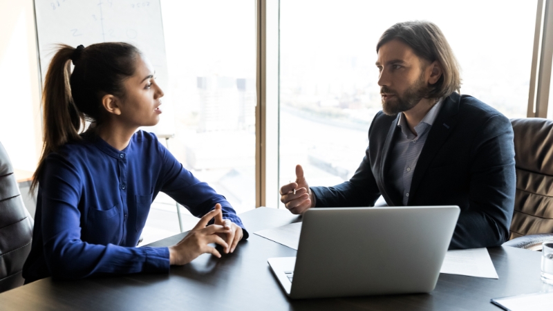 Two colleagues discuss work across a desk with a laptop during a skip-level meeting