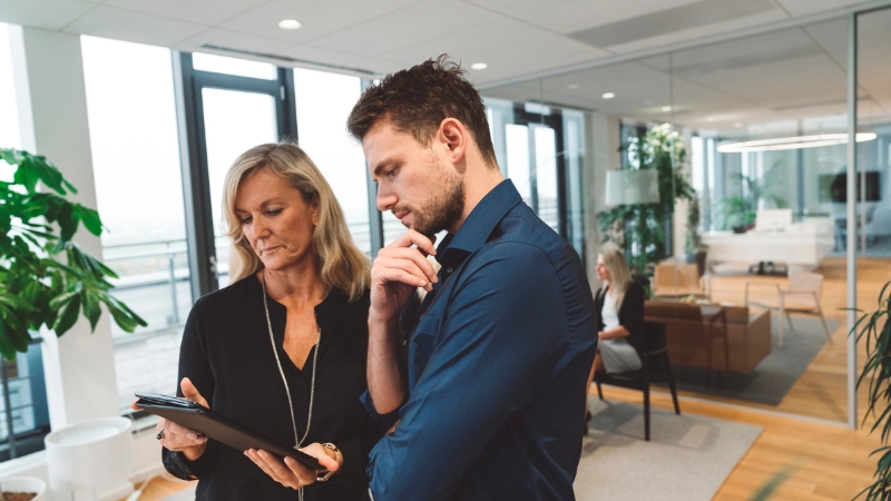 A woman and a man in an office review notes on a tablet while discussing work