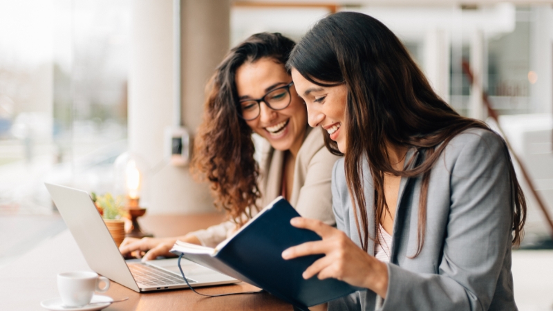 Two women smiling while working together with a laptop and notebook