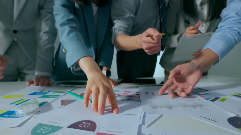 A group of professionals reviews charts and graphs on a table while analyzing factors that influence available market size