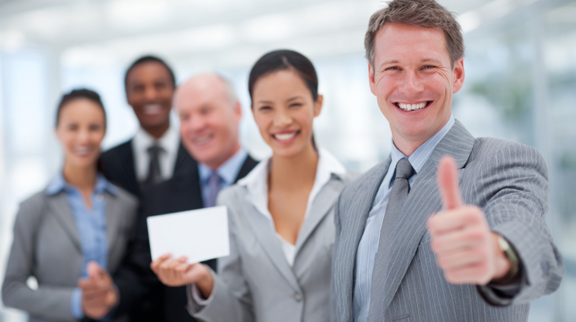 A group of smiling businesspeople in suits stands together, with one man in the foreground giving a thumbs up, symbolizing teamwork and success