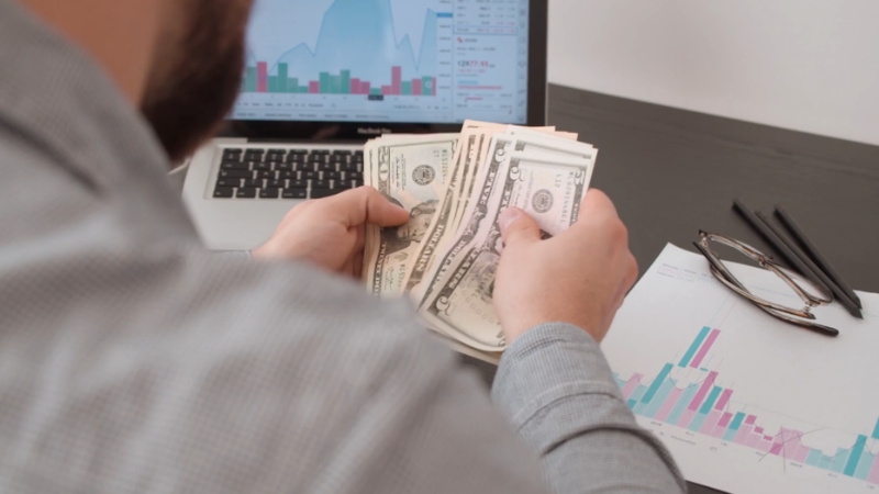 A person counting money at a desk with charts and a laptop showing financial data