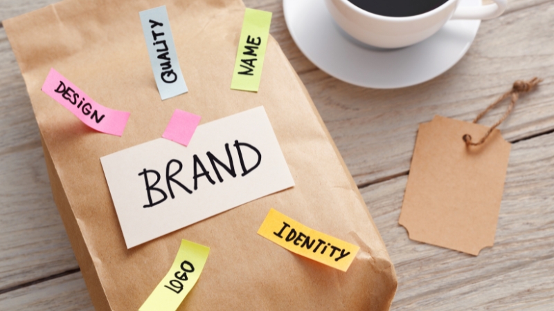 A paper bag labeled “Brand” surrounded by sticky notes with words like design, logo, quality, name, and identity on a wooden table