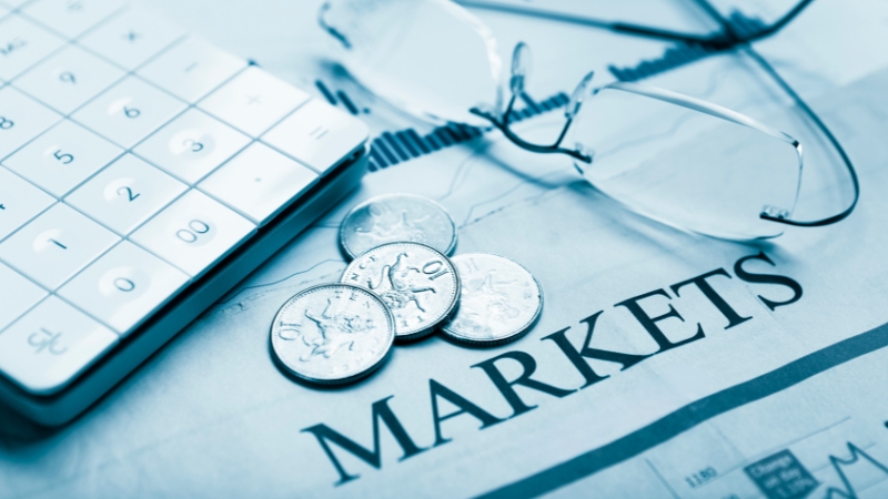 Close-up photo of coins, a calculator, and eyeglasses placed on a financial newspaper page with the word “MARKETS” in focus