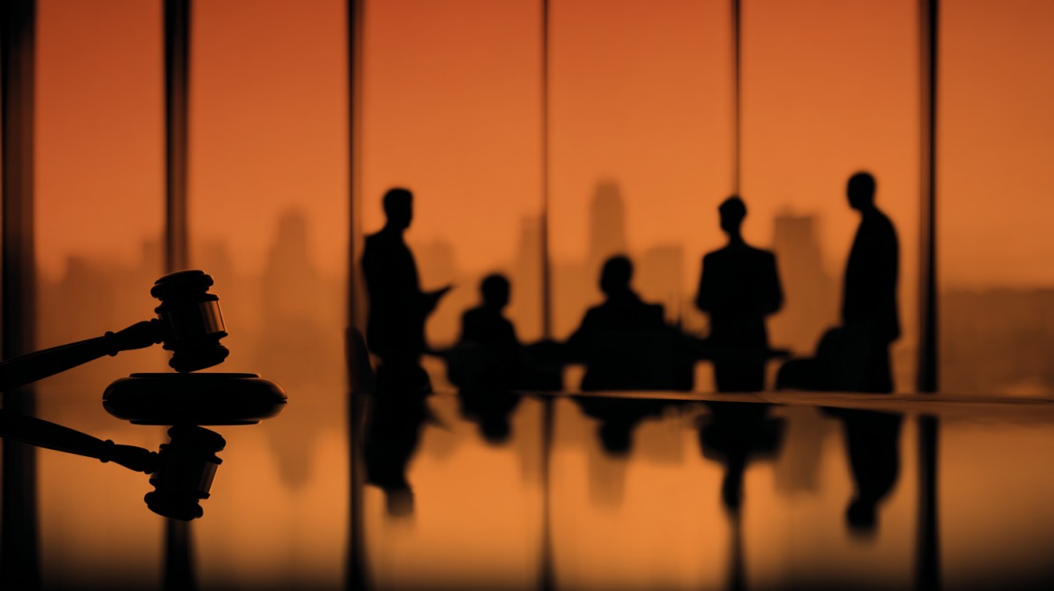 A gavel rests on a conference table with silhouetted businesspeople discussing in front of large windows during sunset, representing legal oversight and compliance integration
