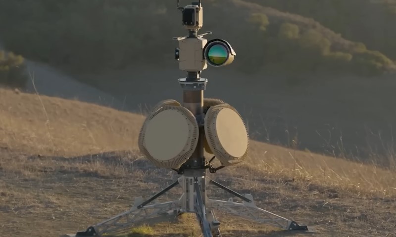 A camera mounted on a tripod stands in the center of a grassy field under a clear blue sky