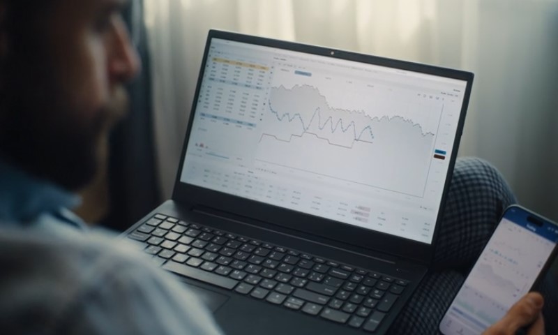 A person examines stock market graphs on a laptop and smartphone, showing fluctuating trends