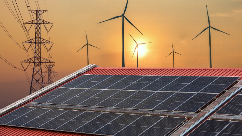 Solar panels on a rooftop with wind turbines and power lines in the background at sunset