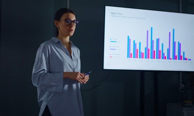 Woman in glasses presenting data on a screen with colorful bar charts labeled "Analytics."