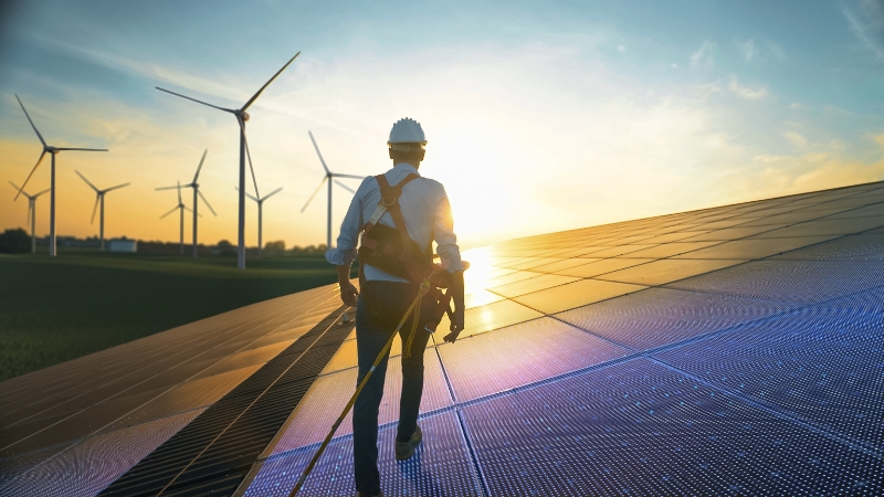 Engineer walking across solar panels with wind turbines in the background at sunset