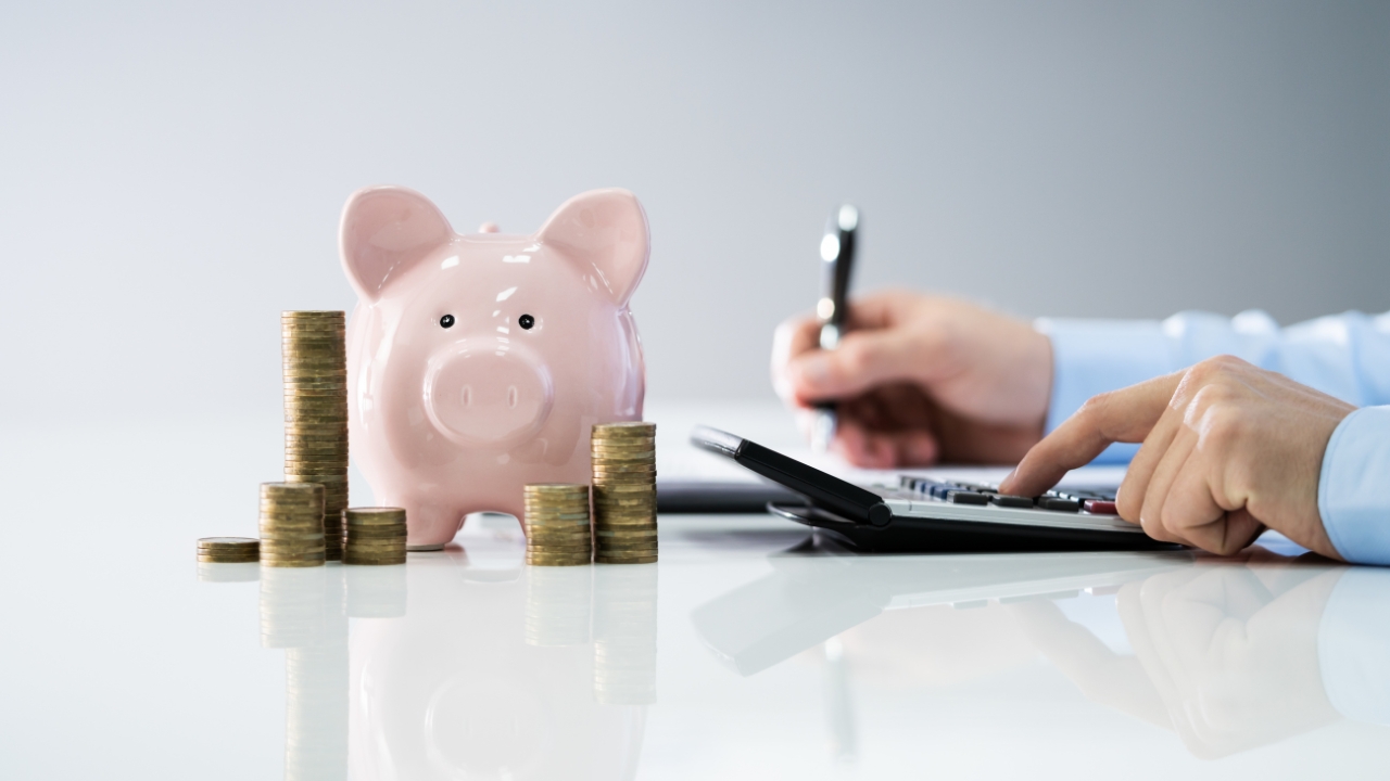 Piggy bank with coin stacks and a person using a calculator to track hidden compliance costs