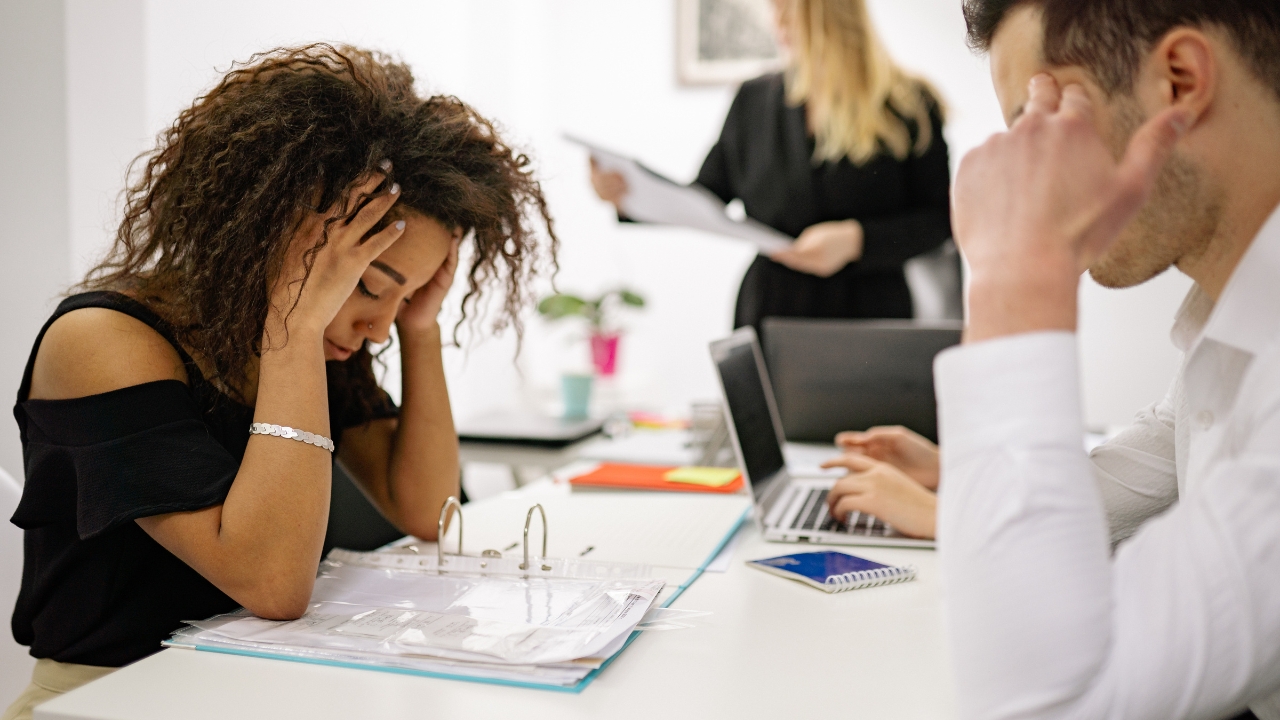 Employees look stressed while reviewing paperwork and working on laptops in an office