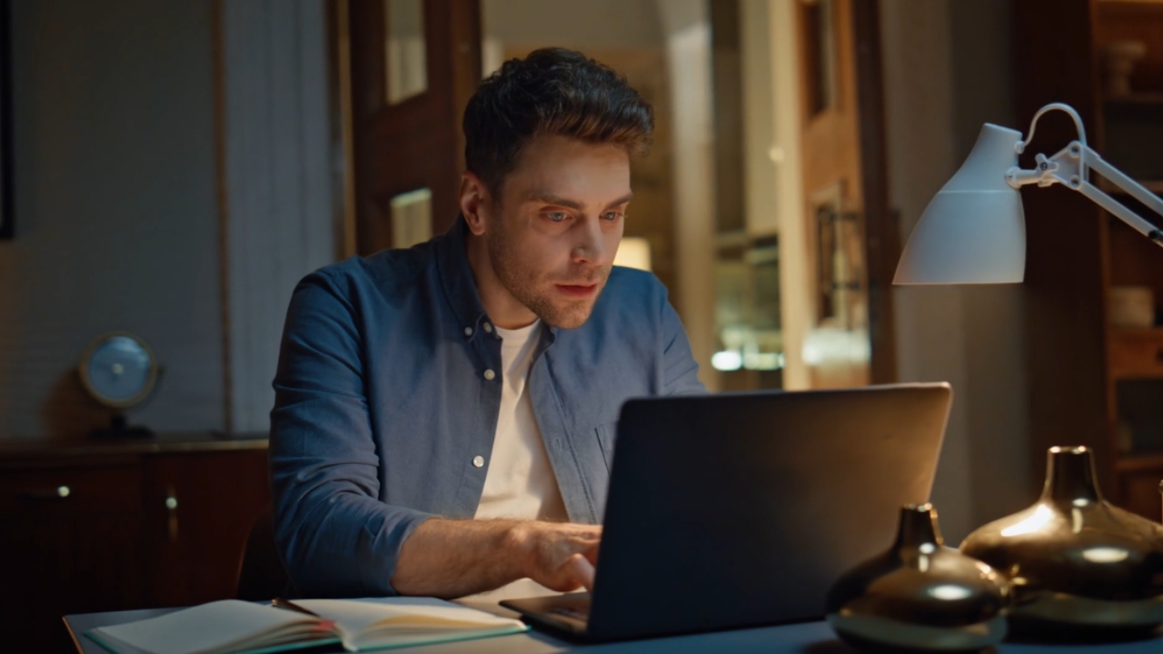Man works on a laptop at home at night, focused on a low-cost remote job