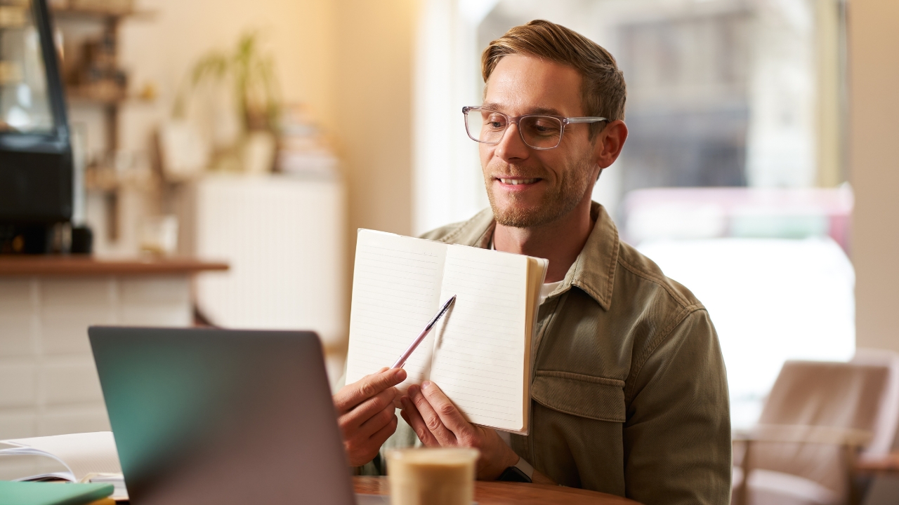 Man teaches online at home, holding a notebook and speaking to a laptop