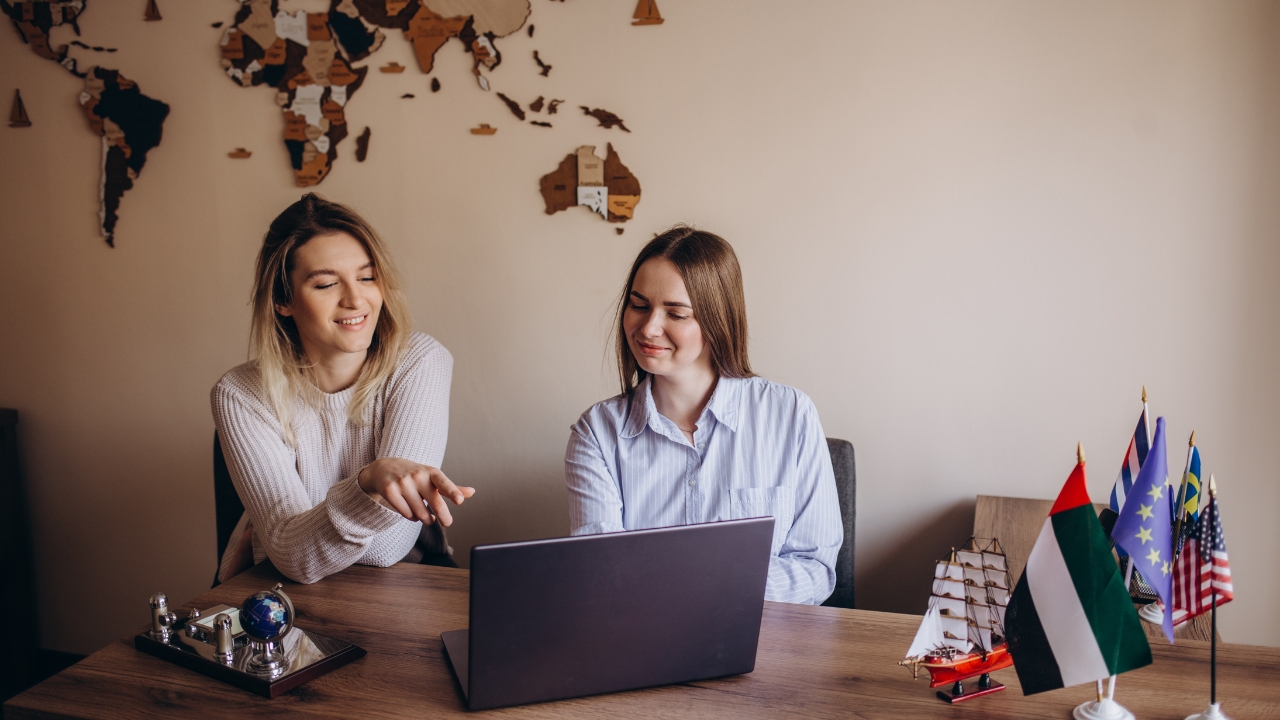 Two women sit at a desk with a laptop, discussing travel plans with a world map on the wall behind them